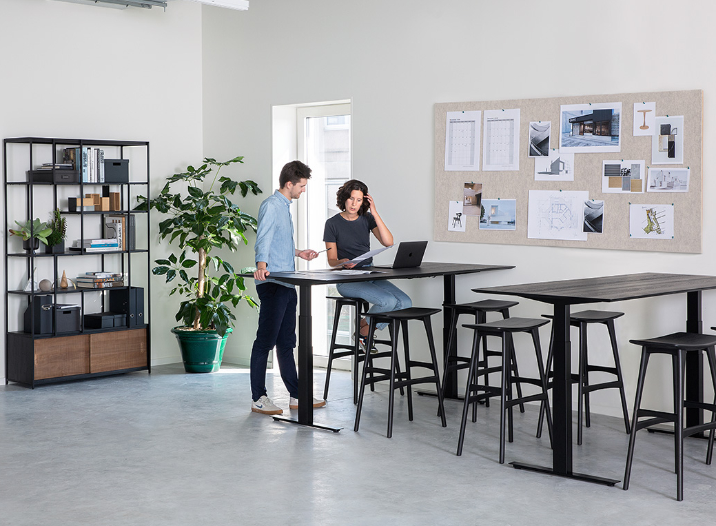 Live Light | Employees interacting in an office space with the Bok Adjustable Desk, Oak Osso Counter Stool and Studio Rack