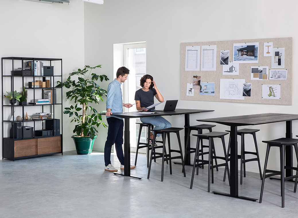Live Light | Employees interacting in an office space with the Bok Adjustable Desk, Oak Osso Counter Stool and Studio Rack Live Light | Employees interacting in an office space with the Bok Adjustable Desk, Oak Osso Counter Stool and Studio Rack